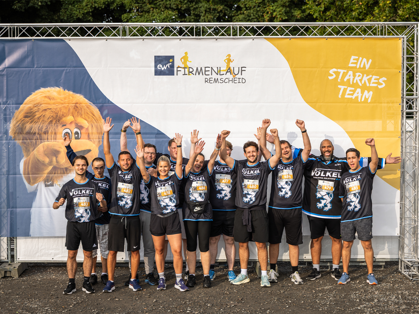 Das VÖLKEL Team jubelt vor dem Banner des Firmenlaufs Remscheid 2025. Ein starkes Team mit voller Energie und Motivation.