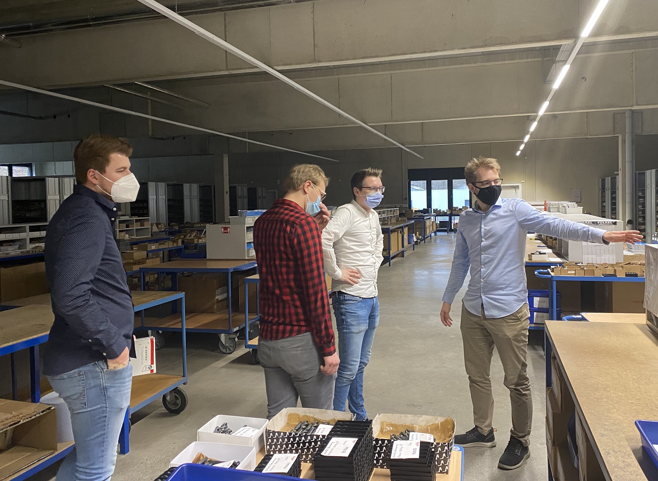 Werksbesichtigung in einer Produktionshalle mit blauen Werkstattwagen, Regalen und Kartons, während ein Mitarbeiter auf einen Bereich zeigt.
