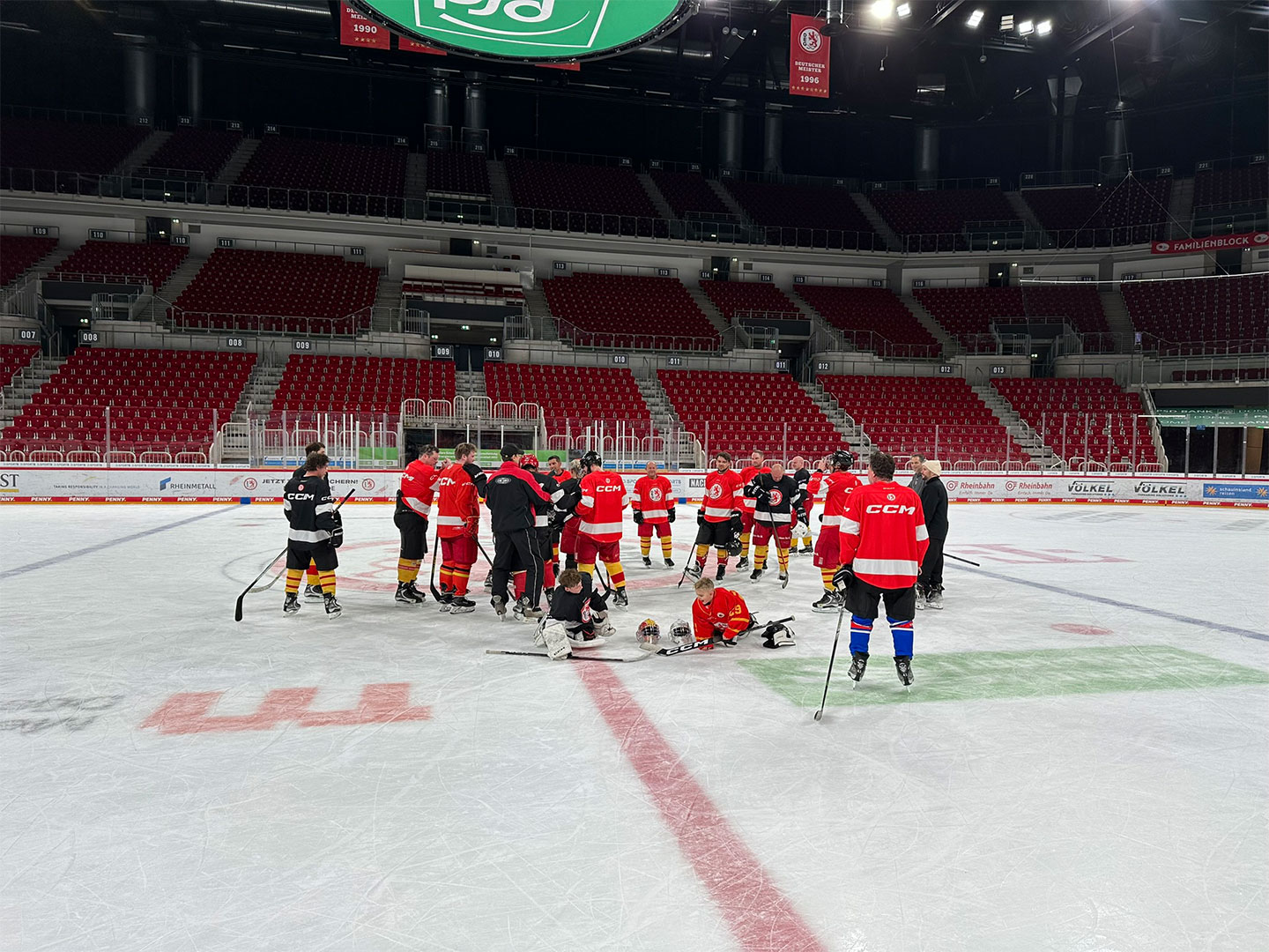 Eine Gruppe von Eishockeyspielern in roten und schwarzen Trikots steht auf dem Eis und erhält Anweisungen.
