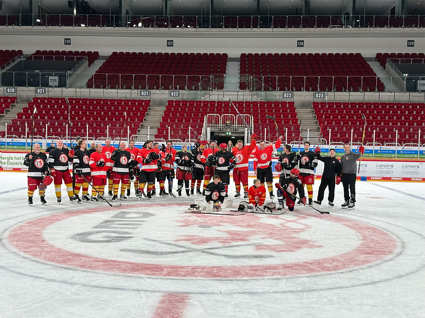 Eine Gruppe von Teilnehmer in roten und schwarzen Trikots posiert auf einer Eisfläche in einem Stadion.