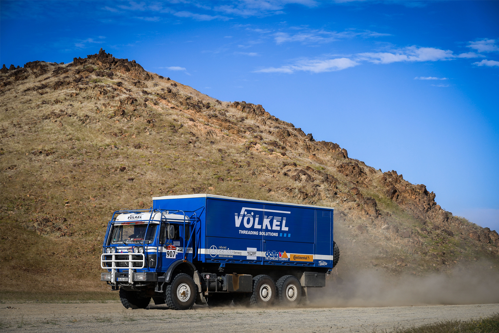 Chemin poussiéreux dans le désert avec un camion sponsorisé par Völkel et montagnes en arrière-plan au Rallye Dakar.