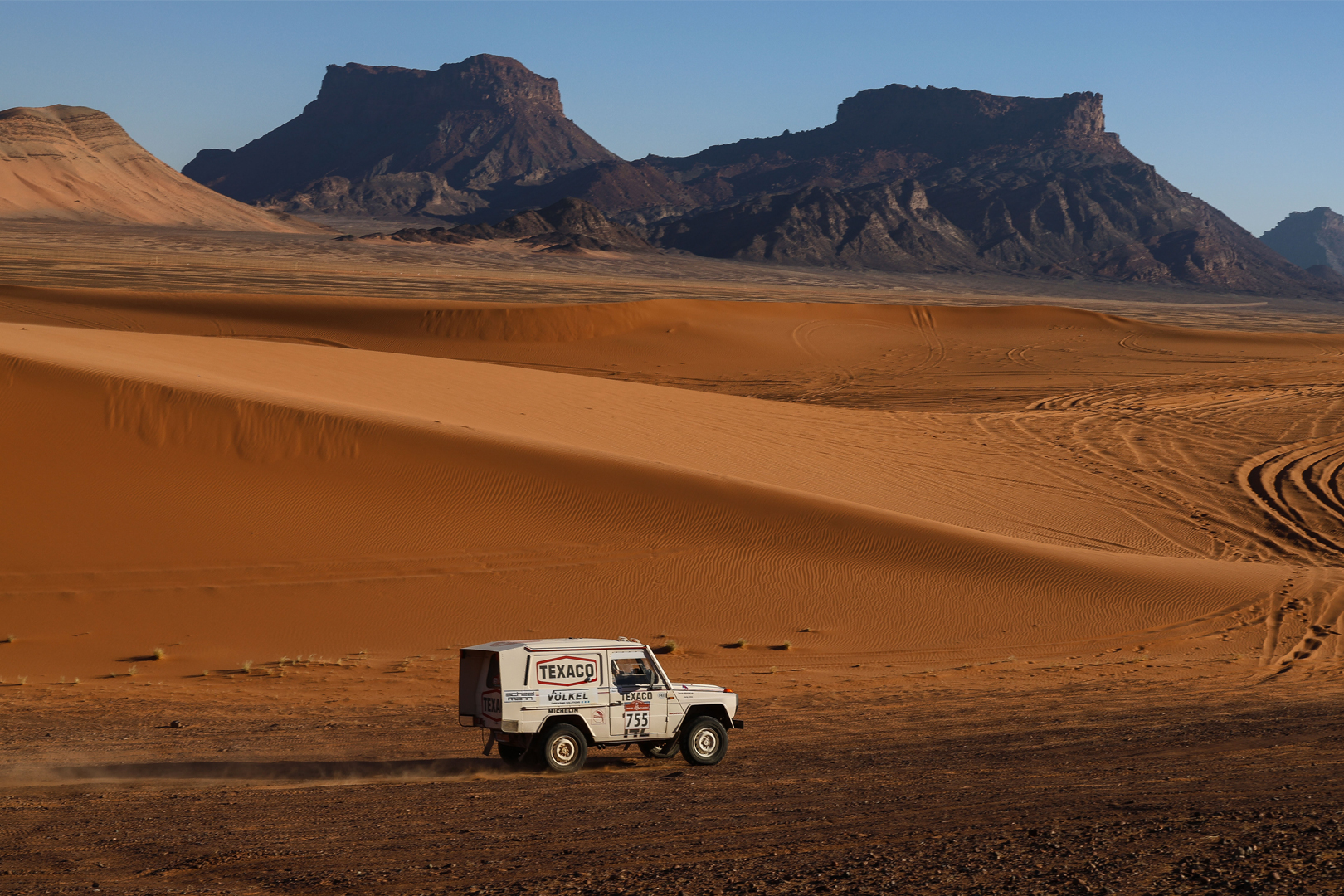 Rallye-raid dans le désert avec voiture Texaco et VÖLKEL projetant du sable.