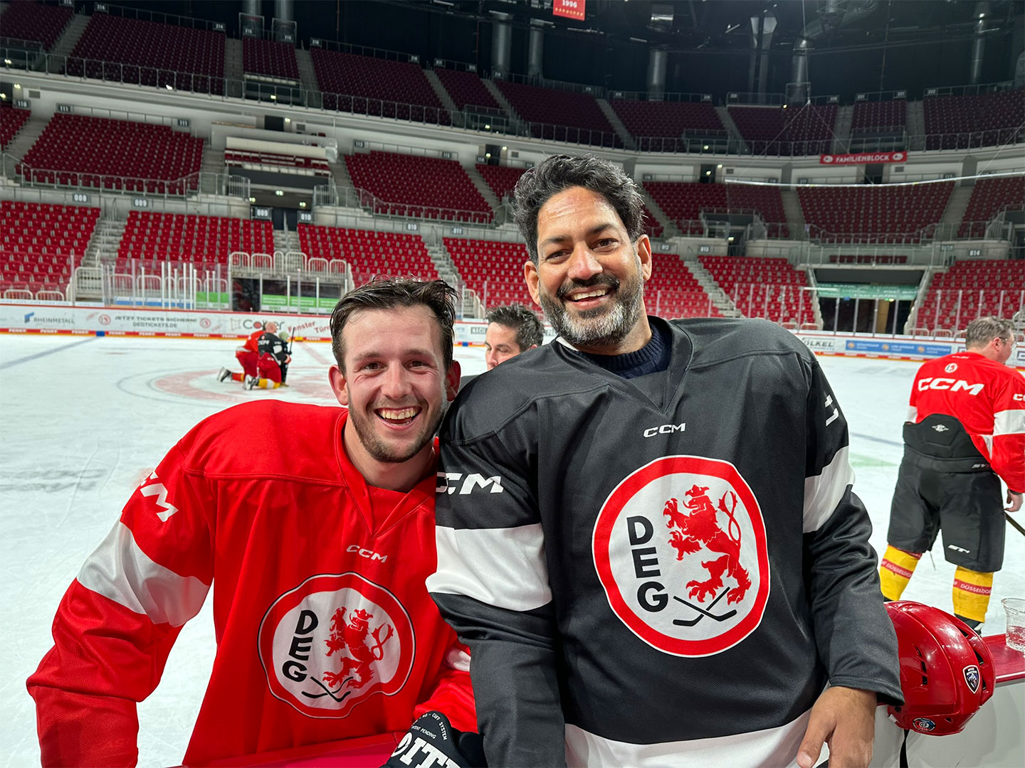 Zwei Eishockeyspieler auf einer Eisbahn. Der linke trägt ein rotes Trikot, der rechte ein schwarzes.