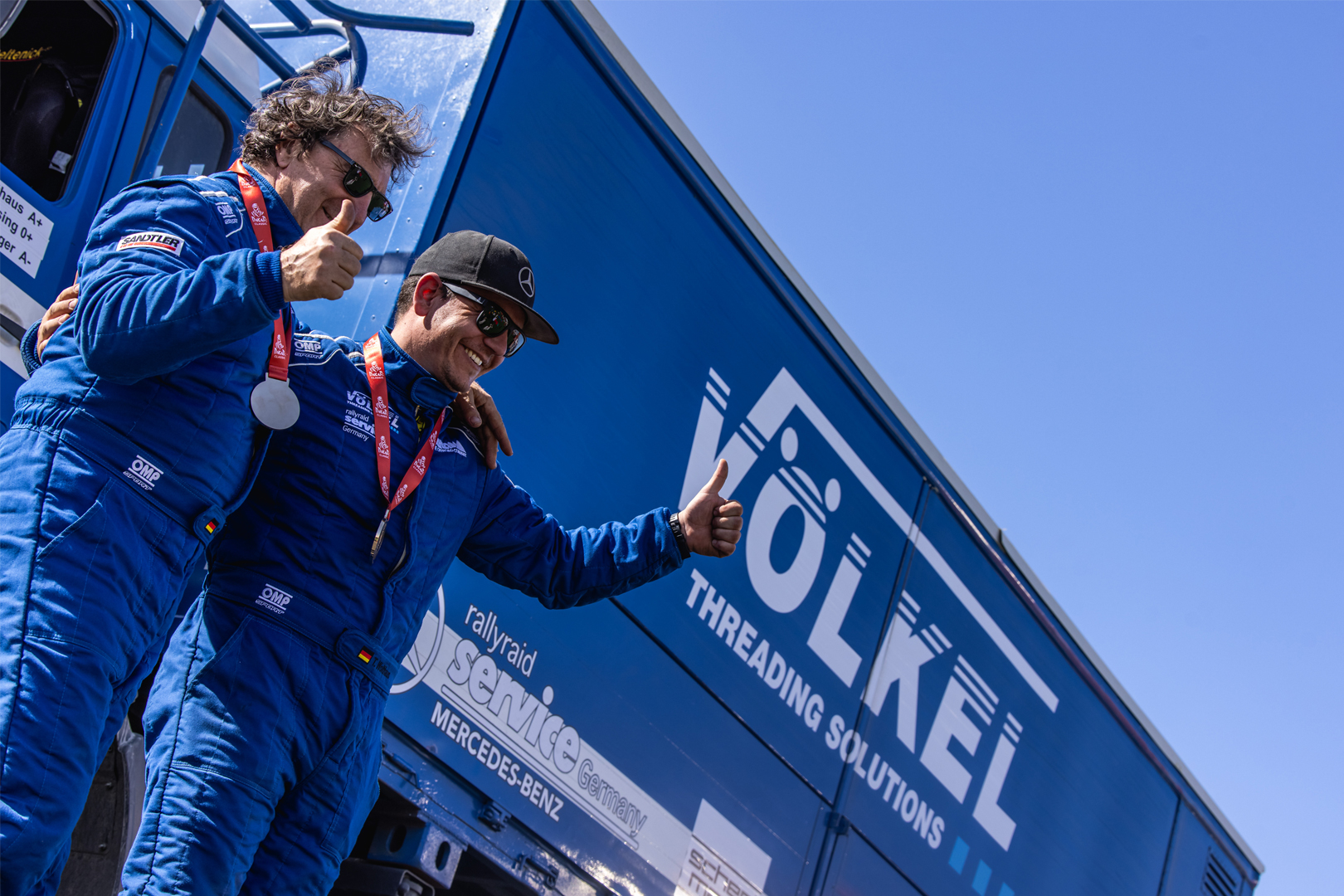 Deux personnes en combinaison de course bleue devant un camion VÖLKEL au Rallye Dakar.