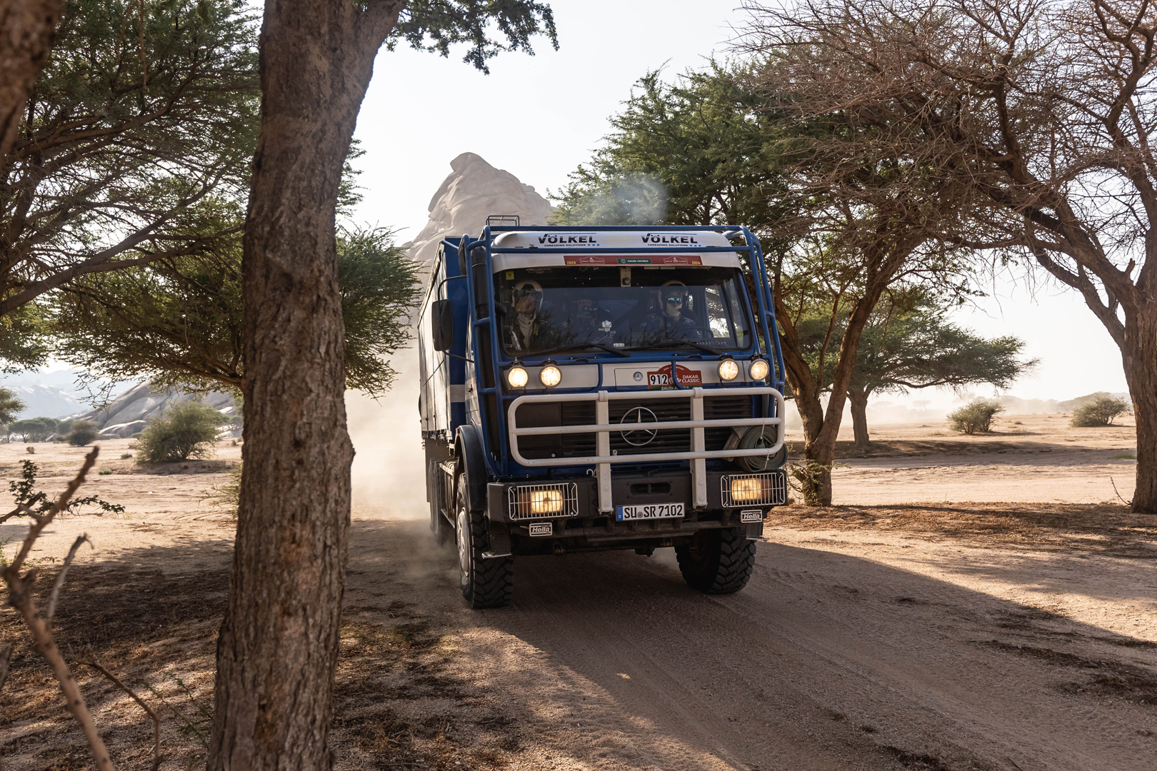 Un camion bleu de Völkel roulant à travers un paysage désertique poussiéreux au Rallye Dakar, montagnes en arrière-plan.