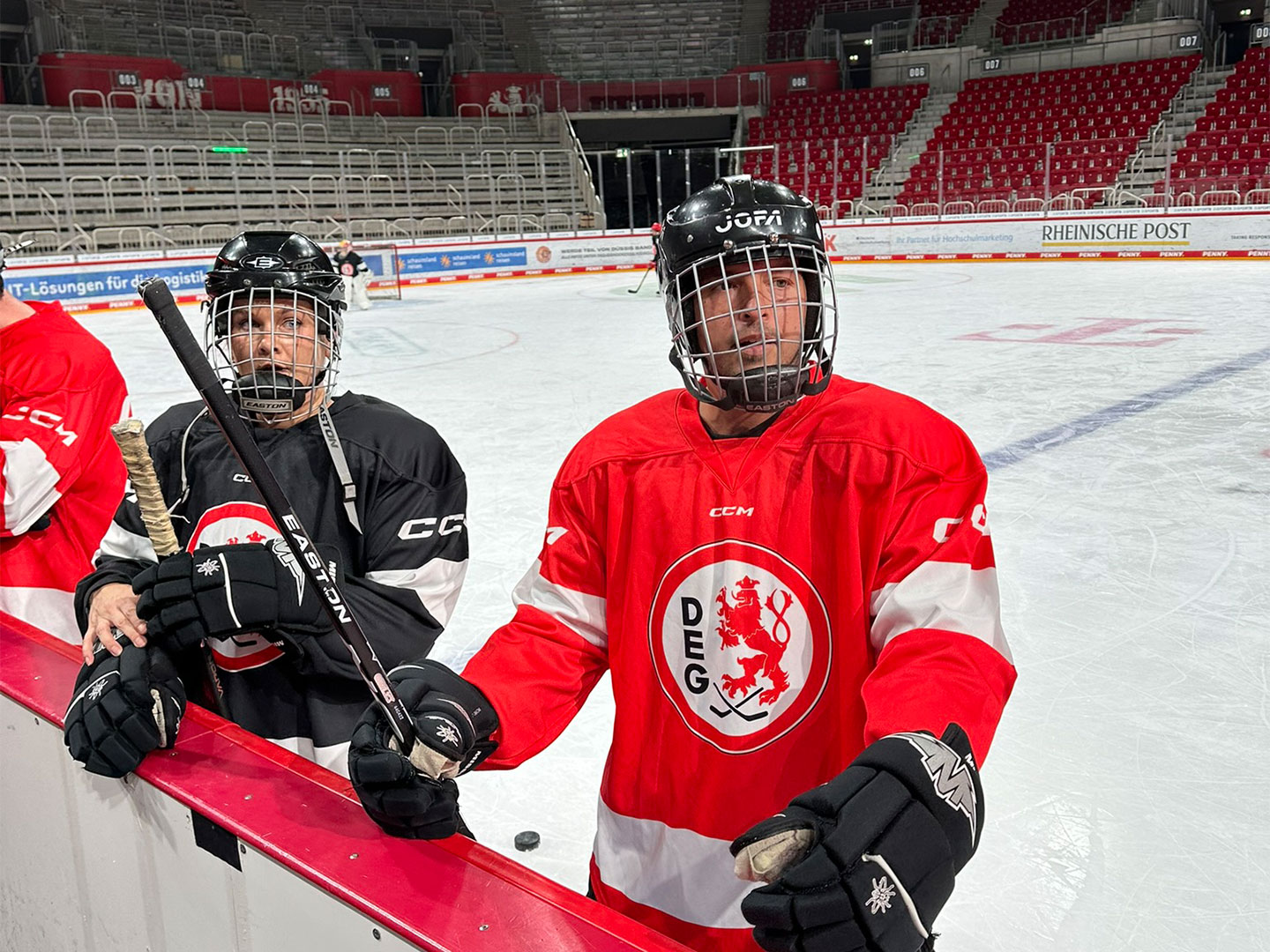 Zwei Eishockeyspieler, einer in schwarzem Trikot und einer in rotem DEG-Trikot, stehen am Spielfeldrand."