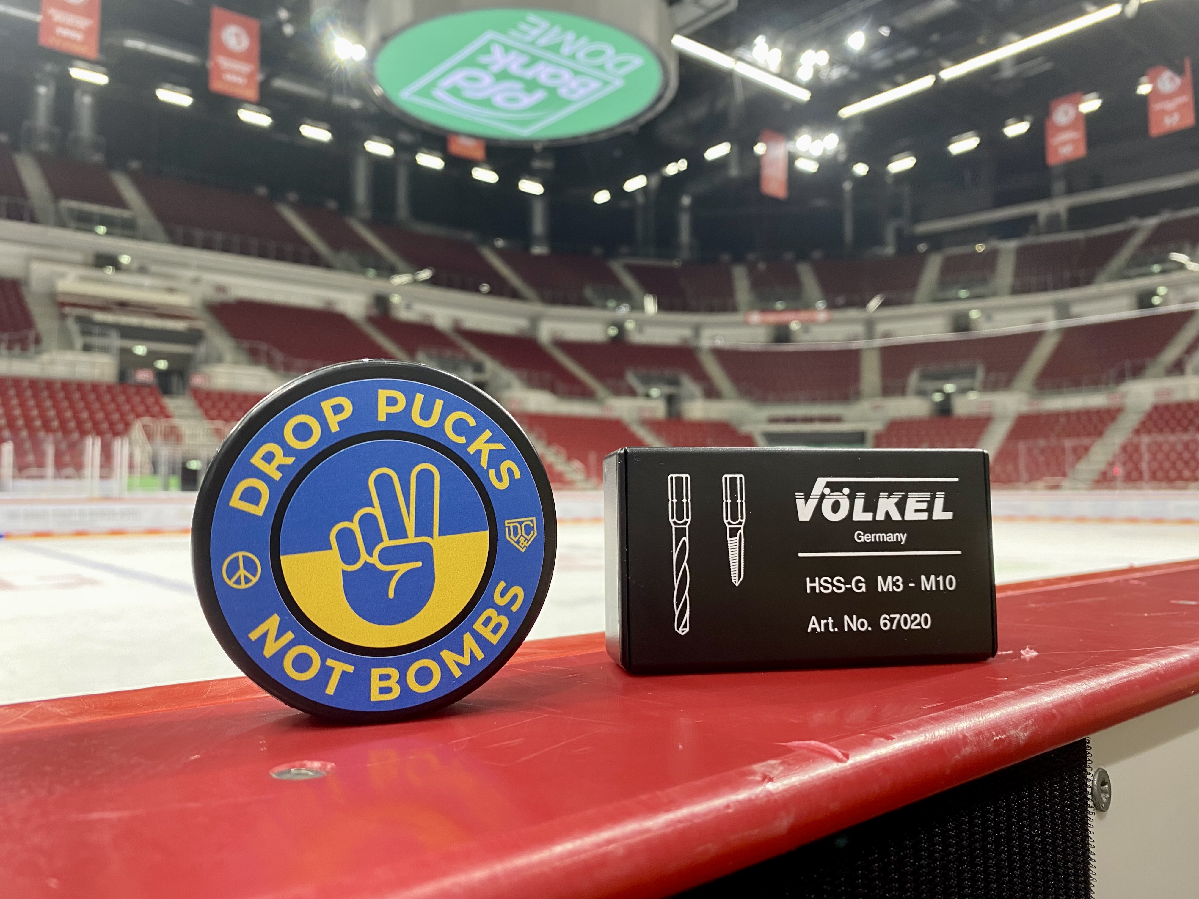 Eishockey-Puck mit Aufdruck „Drop Pucks Not Bombs“ in ukrainischen Farben neben einem VÖLKEL Gewindebohrer-Set in einer Eishockey-Arena