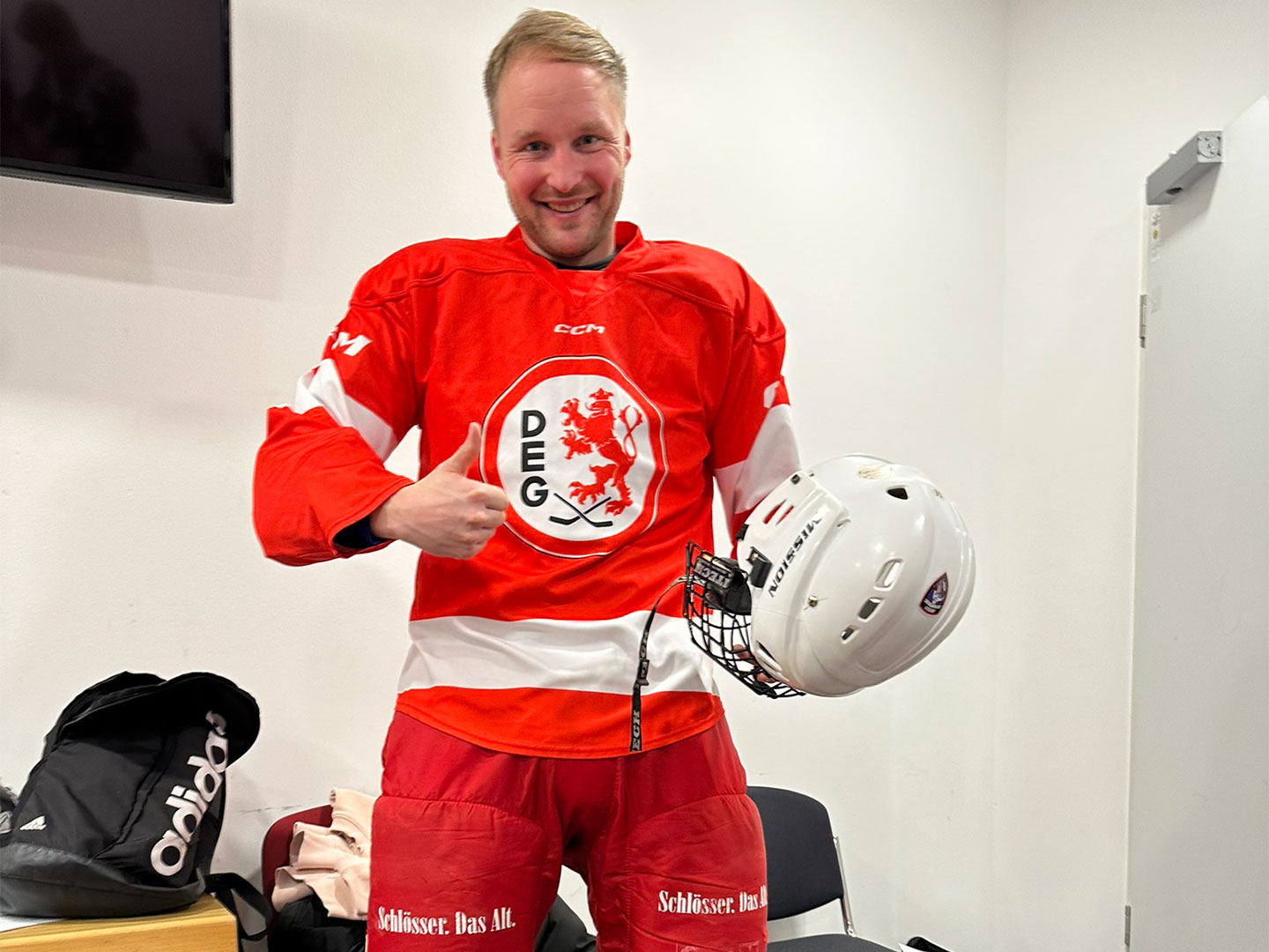 Eine Person in Eishockeyausrüstung mit ein rotes Trikot, hält ein Helm in der Hand und steht in einer Umkleidekabine.