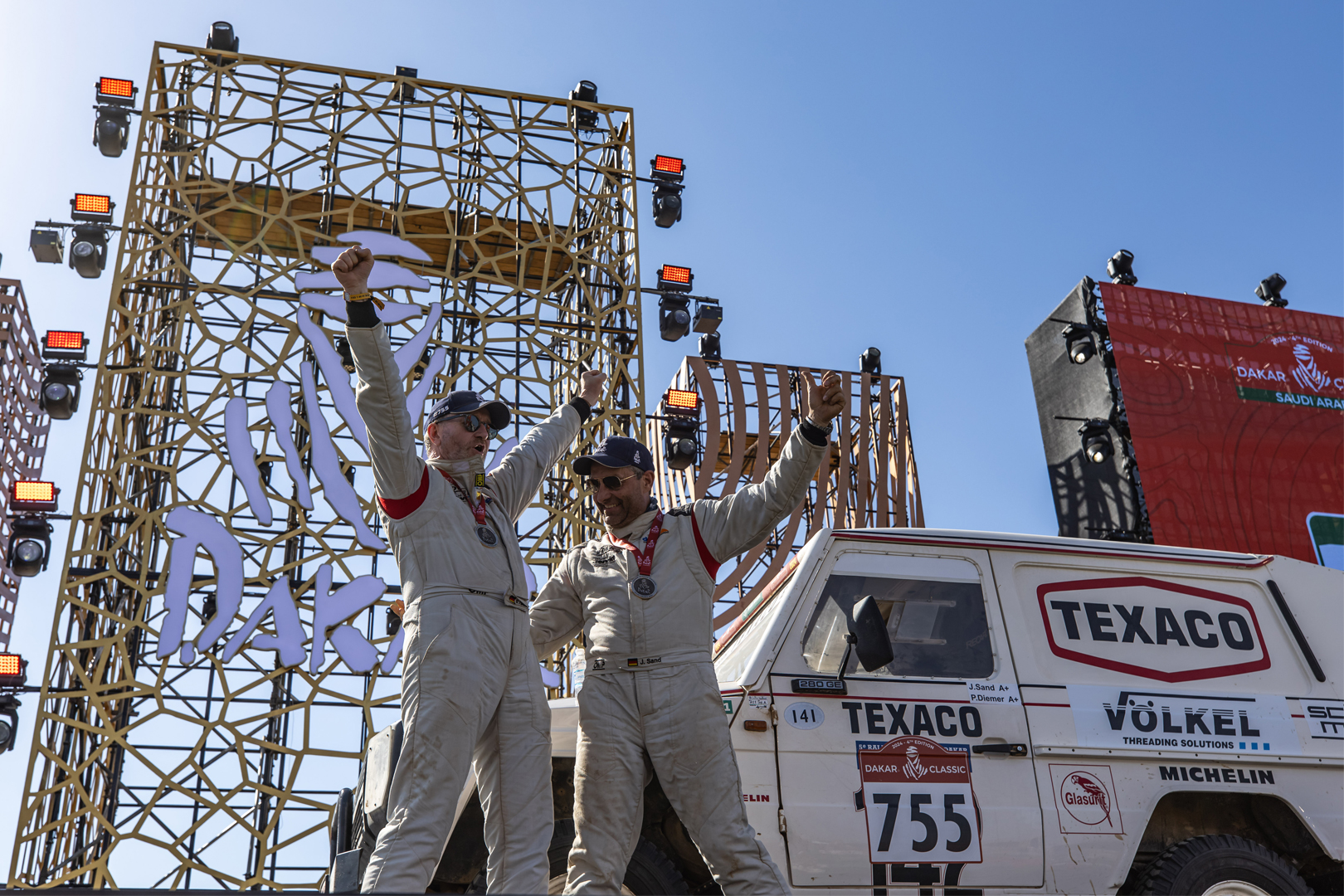 Deux personnes en combinaison de course célébrant sur un véhicule avec le numéro 755 et le logo VÖLKEL au Rallye Dakar.
