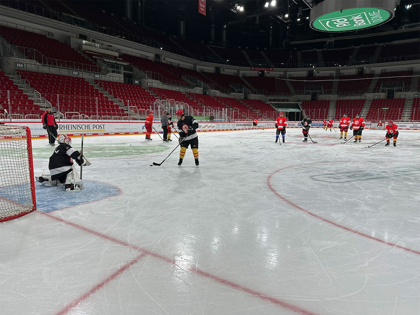 Ein Eishockeytorwart in schwarzer Ausrüstung steht vor dem Tor, während Spieler in roten und schwarzen Trikots auf dem Eis sind.