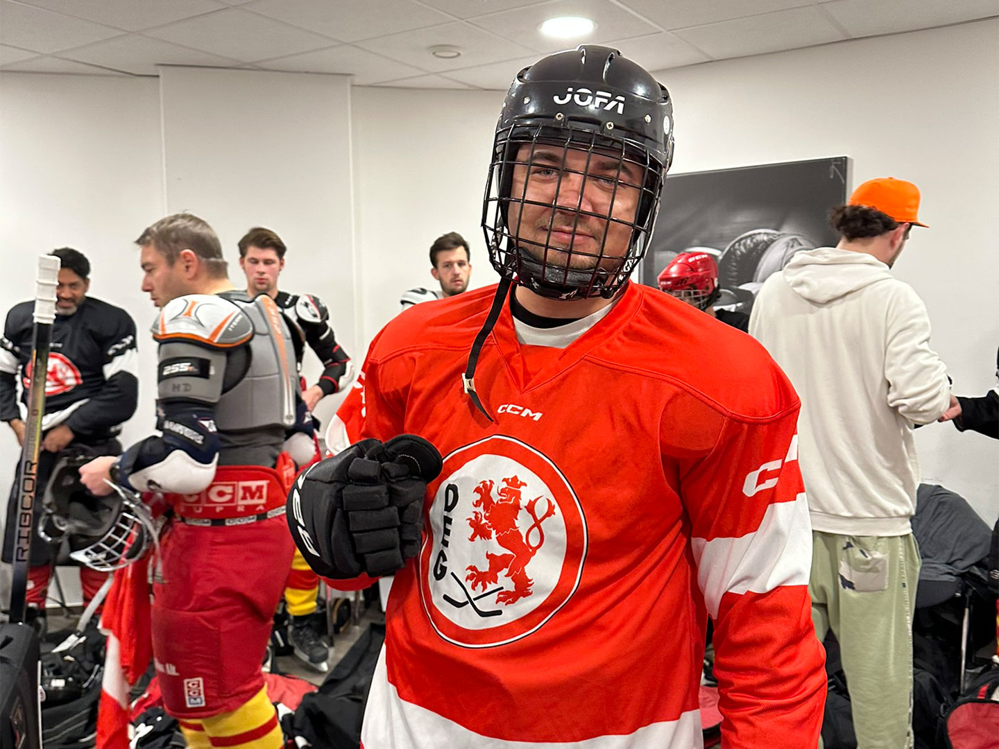 Eine Person in Eishockeyausrüstung mit ein rotes Trikot, mit aufgesetzten Helm steht in einer Umkleidekabine.
