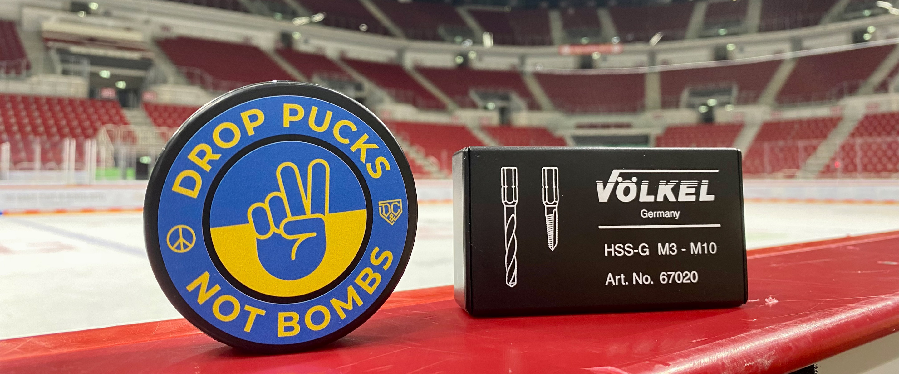 Eishockey-Puck mit Aufschrift „Drop Pucks Not Bombs“ in Blau-Gelb neben VÖLKEL Gewindebohrer-Set