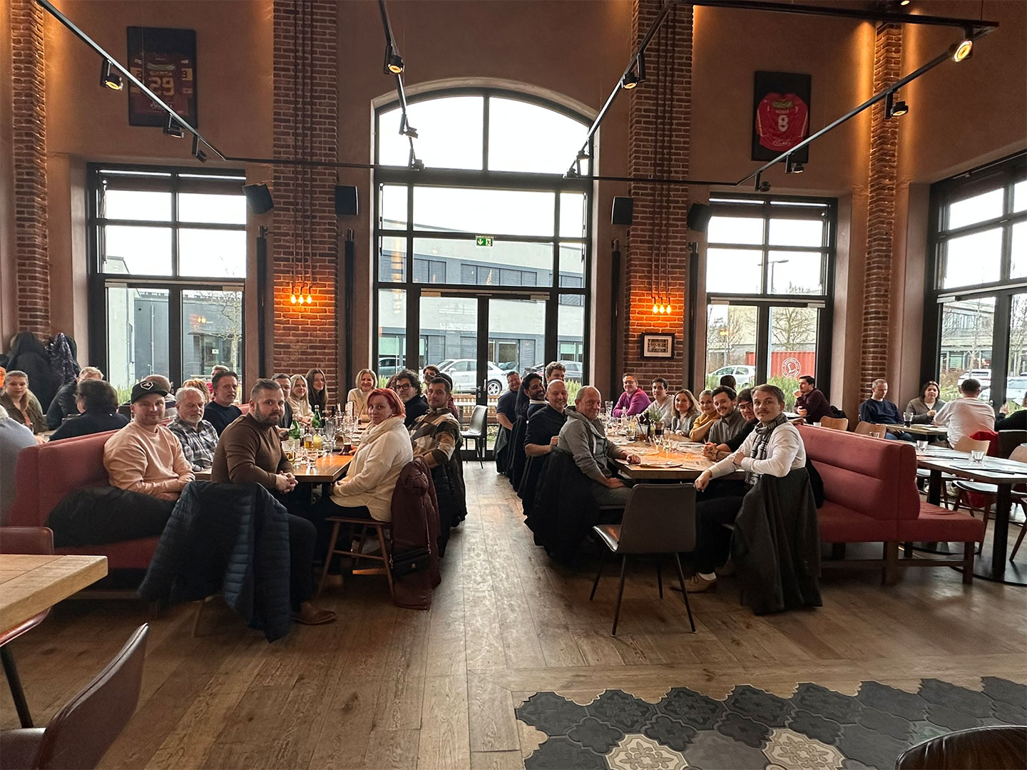 Eine Gruppe von Menschen sitzt an Tischen in einem Restaurant mit großen Fenstern und Backsteinwänden.