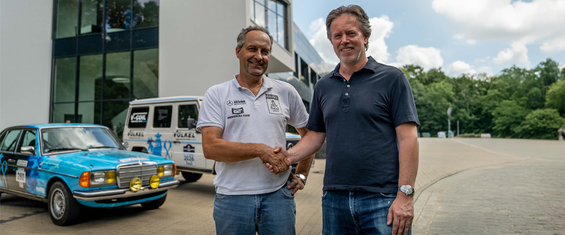 Handschlag zur Partnerschaft im Rally Raid vor dem blauen Rennfahrzeug am VÖLKEL Firmensitz