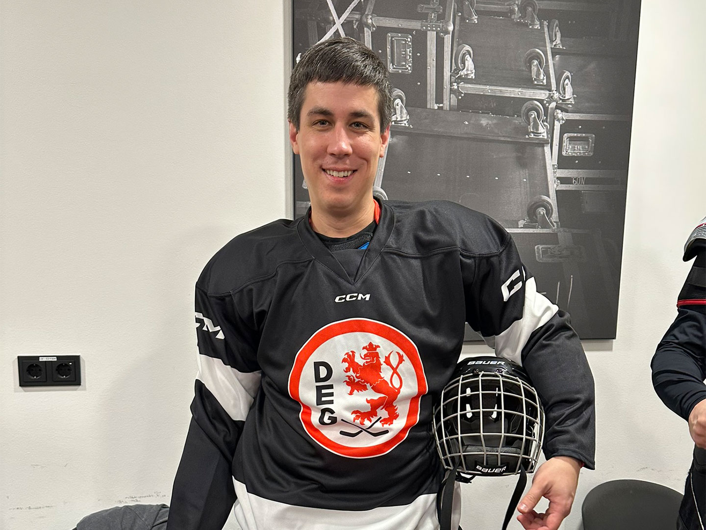 Eine Person in Eishockeyausrüstung mit ein schwarzes Trikot, hält ein Helm unter sein Arm und steht in einer Umkleidekabine.