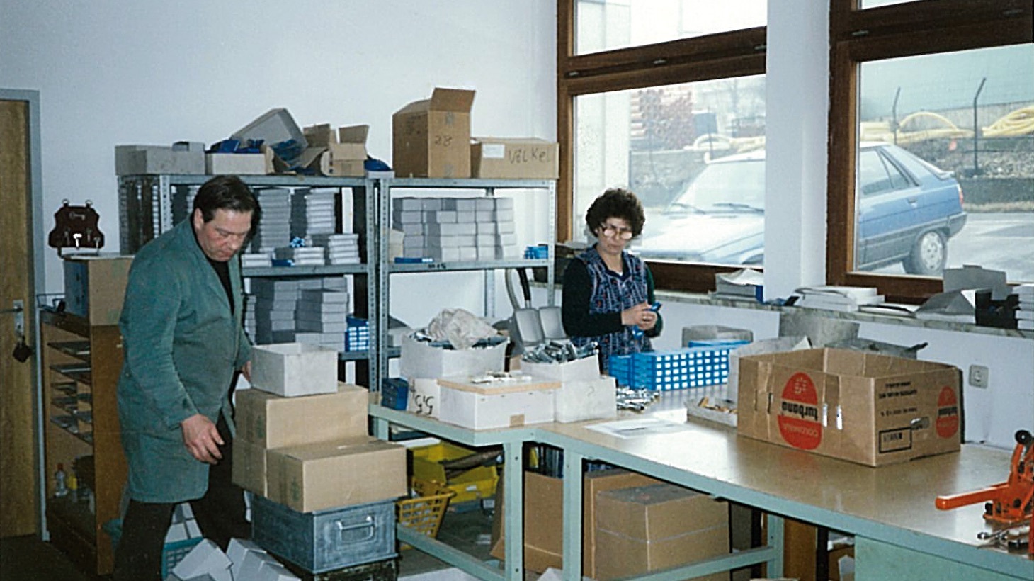 Two individuals working in a room with shelves full of boxes and files, a blue car visible through the window.