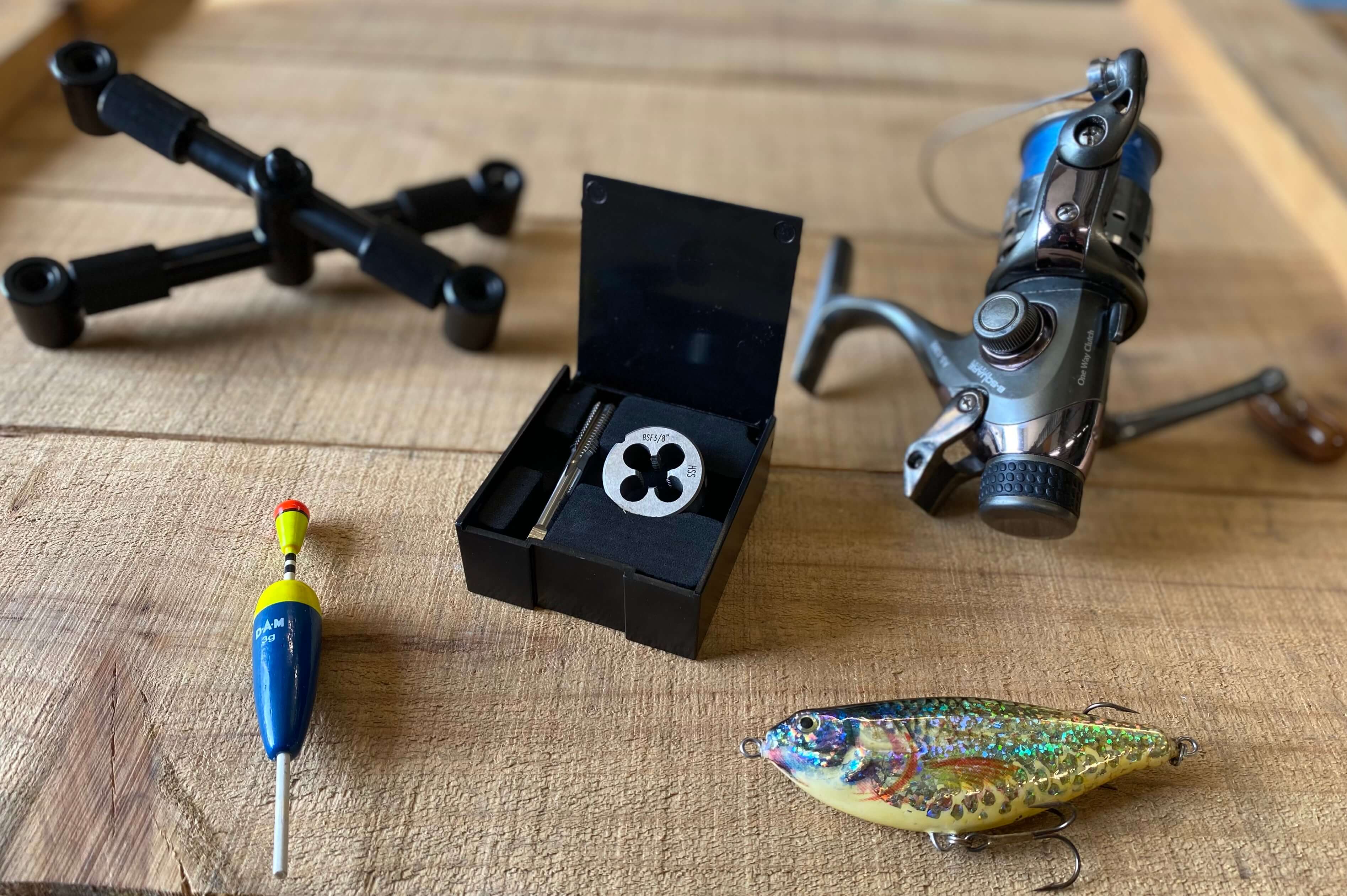 Gewindeschneidwerkzeug in Box auf Holzoberfläche neben Angelrolle, Köder und Zubehör, dargestellt in einer Werkstatt- und Anwendungsszene.