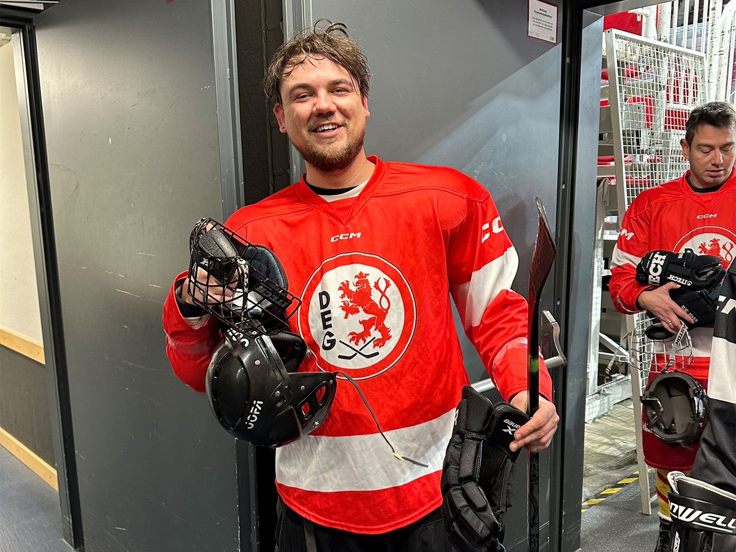Ein Teilnehmer trägt ein rotes Eishockeytrikot mit dem Logo "DEG" und hält einen schwarzen Helm und einen Eishockeyschläger.