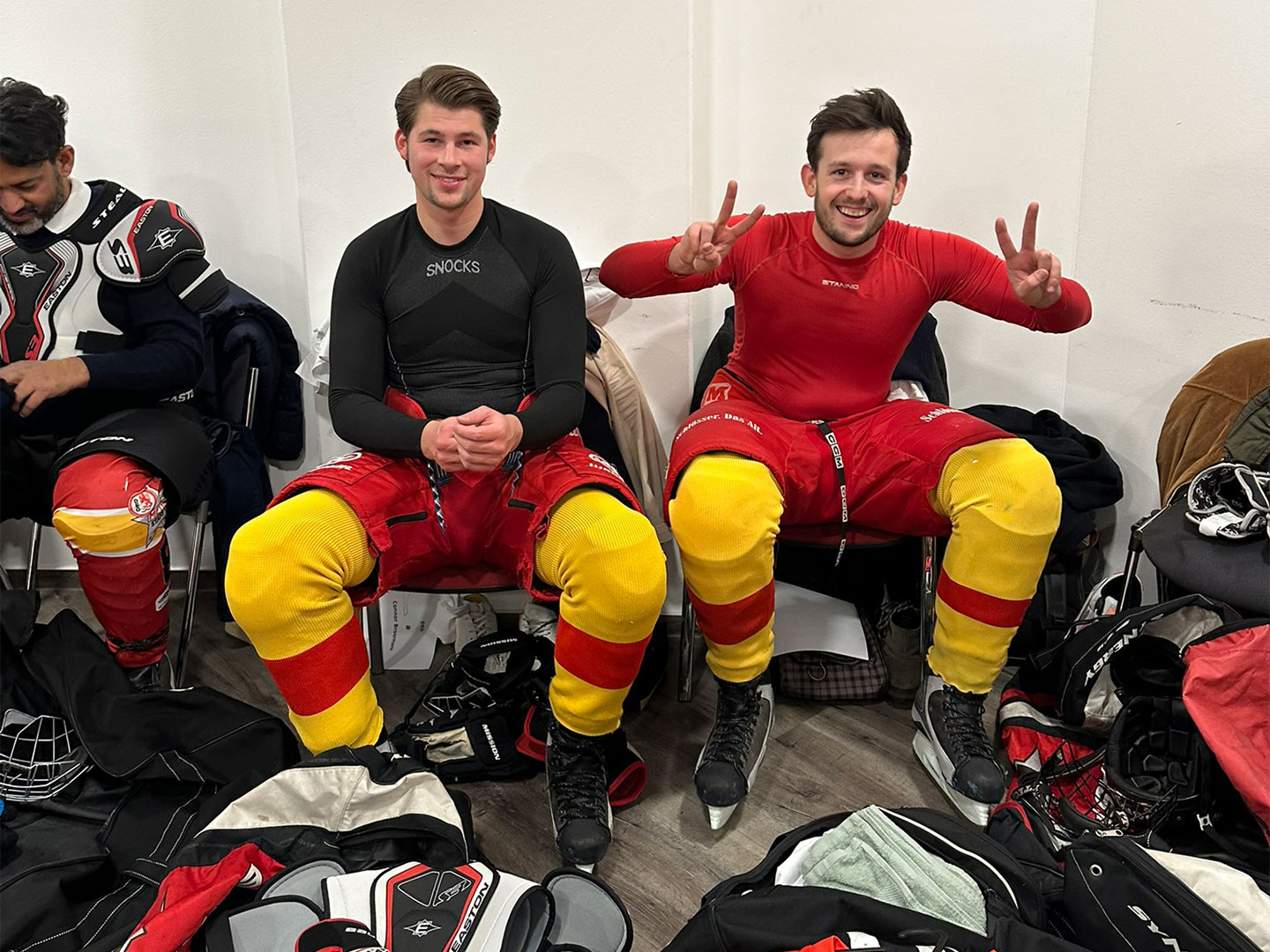 Zwei Personen in Eishockeyausrüstung sitzen in einer Umkleidekabine.