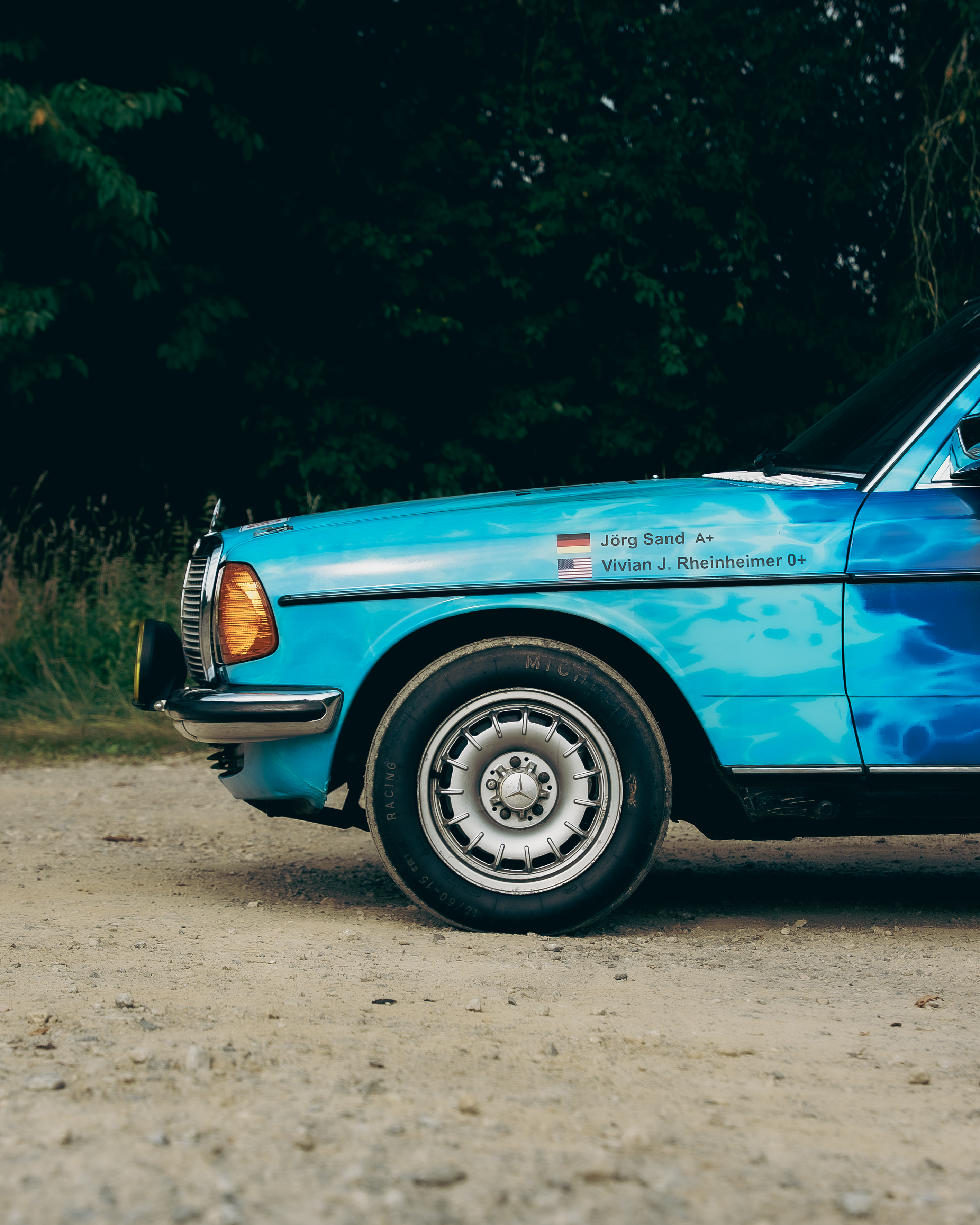 Mercedes-Benz 280E W123 Radseite Sponsoren Seitenansicht mit Fokus auf Vorderrad und Sponsorenaufdrucke am Mercedes-Benz 280E W123, Carrera Panamericana.