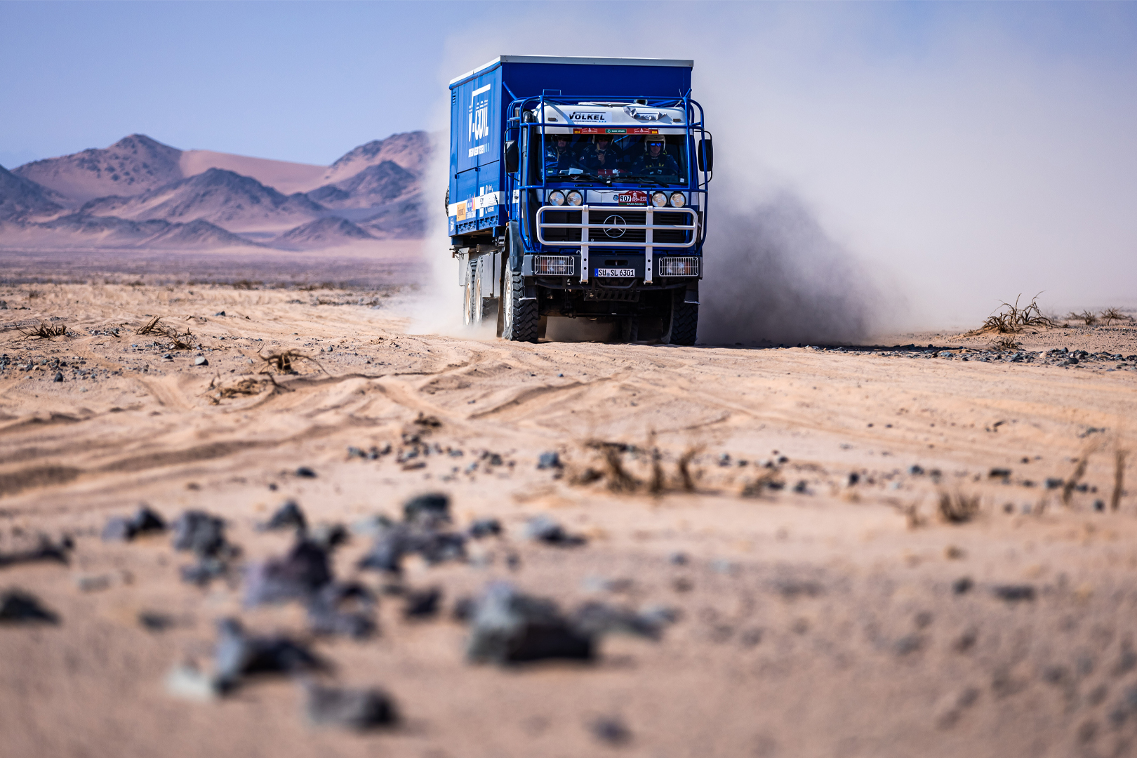 Camion bleu de Völkel soulevant la poussière tout en roulant dans le désert au Rallye Dakar, montagnes en arrière-plan.